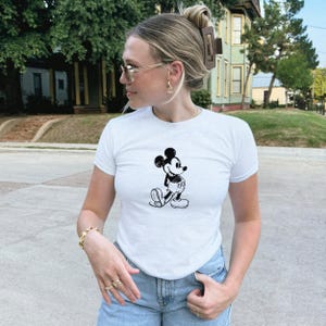 May include: White short-sleeve t-shirt featuring a black and white illustration of Mickey Mouse. The shirt is paired with blue jeans. The image shows a person wearing the shirt outdoors.