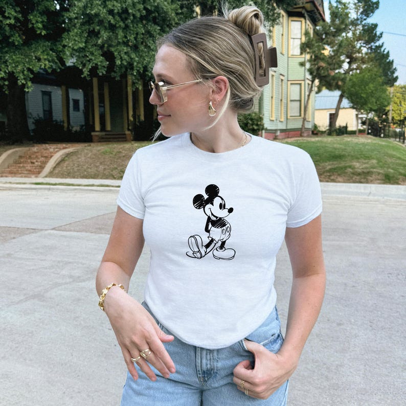 May include: White short-sleeve t-shirt featuring a black and white illustration of Mickey Mouse. The shirt is paired with blue jeans. The image shows a person wearing the shirt outdoors.