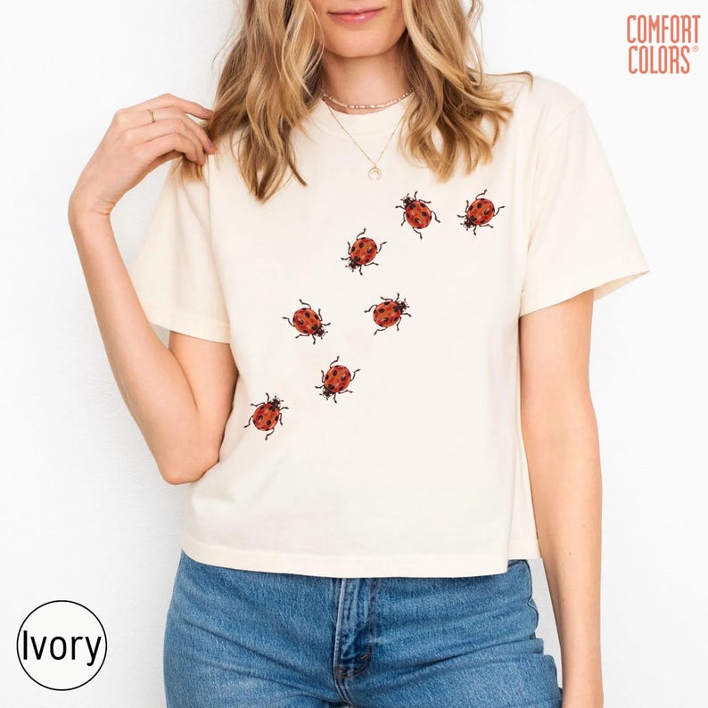 May include: An ivory short-sleeve t-shirt featuring a graphic print of seven small red ladybugs with black spots scattered across the front. The shirt has a crew neckline and is paired with blue denim jeans. The text COMFORT COLORS is visible.