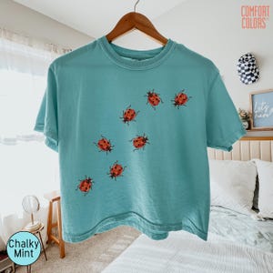 May include: A short-sleeve t-shirt in a chalky mint color featuring a graphic print of seven red ladybugs with black spots scattered across the front. The shirt has a crew neckline and is displayed on a wooden hanger against a light background.