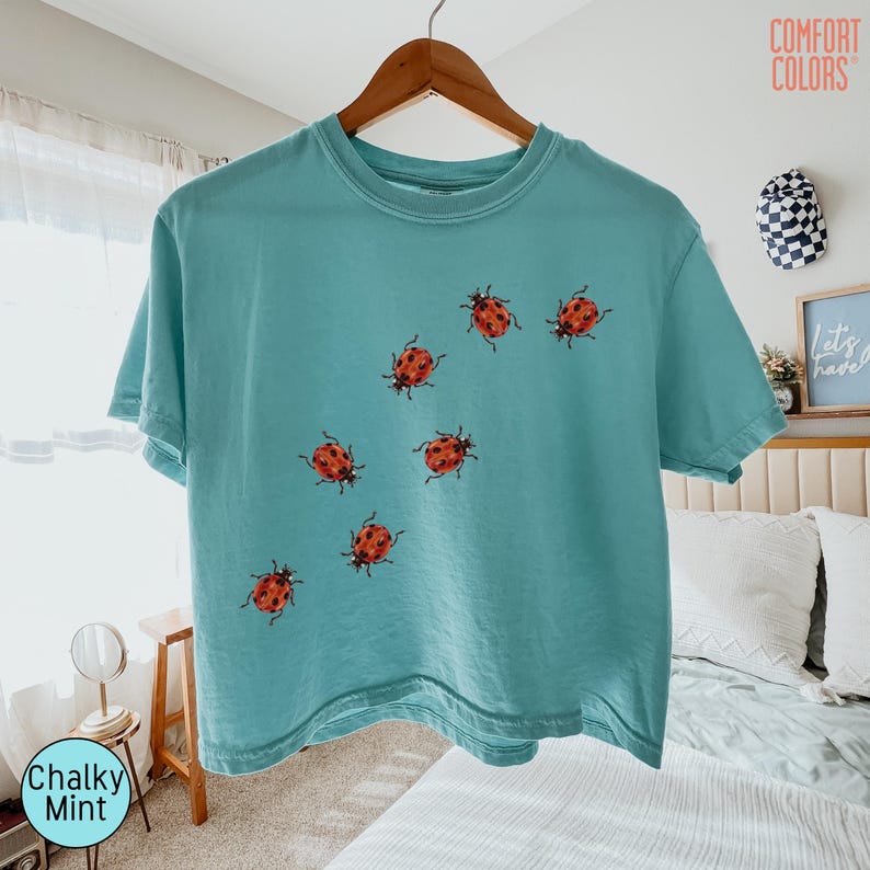 May include: A short-sleeve t-shirt in a chalky mint color featuring a graphic print of seven red ladybugs with black spots scattered across the front. The shirt has a crew neckline and is displayed on a wooden hanger against a light background.