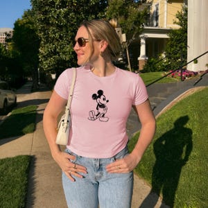 May include: A light pink t-shirt featuring a black graphic of Mickey Mouse. The shirt has short sleeves and a crew neck. The person is wearing blue jeans and sunglasses. A white purse is over the shoulder.
