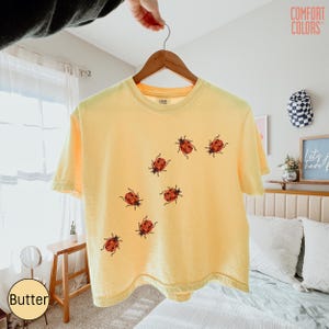 May include: A pale yellow short-sleeve t-shirt featuring a scattered pattern of small red and black ladybug illustrations across the front. The shirt has a crew neckline and is displayed on a wooden hanger against a bright indoor setting.