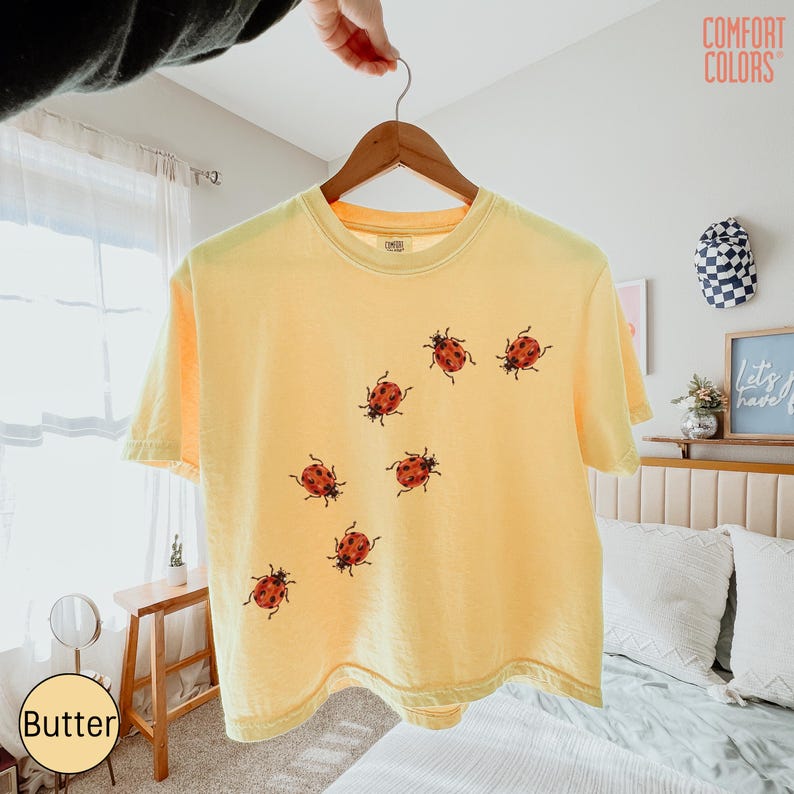May include: A pale yellow short-sleeve t-shirt featuring a scattered pattern of small red and black ladybug illustrations across the front. The shirt has a crew neckline and is displayed on a wooden hanger against a bright indoor setting.