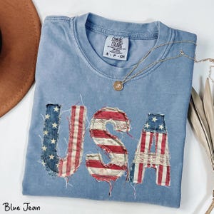 May include: A blue jean colored t-shirt with the word "USA" appliqued on the front in a patriotic design. The letters are made of denim and red and white striped fabric. A gold necklace with a pendant rests on the shirt.