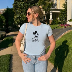 May include: Light blue t-shirt featuring a black and white illustration of Mickey Mouse. The shirt has short sleeves and a crew neck. The person is wearing blue jeans and sunglasses, and carrying a white purse.