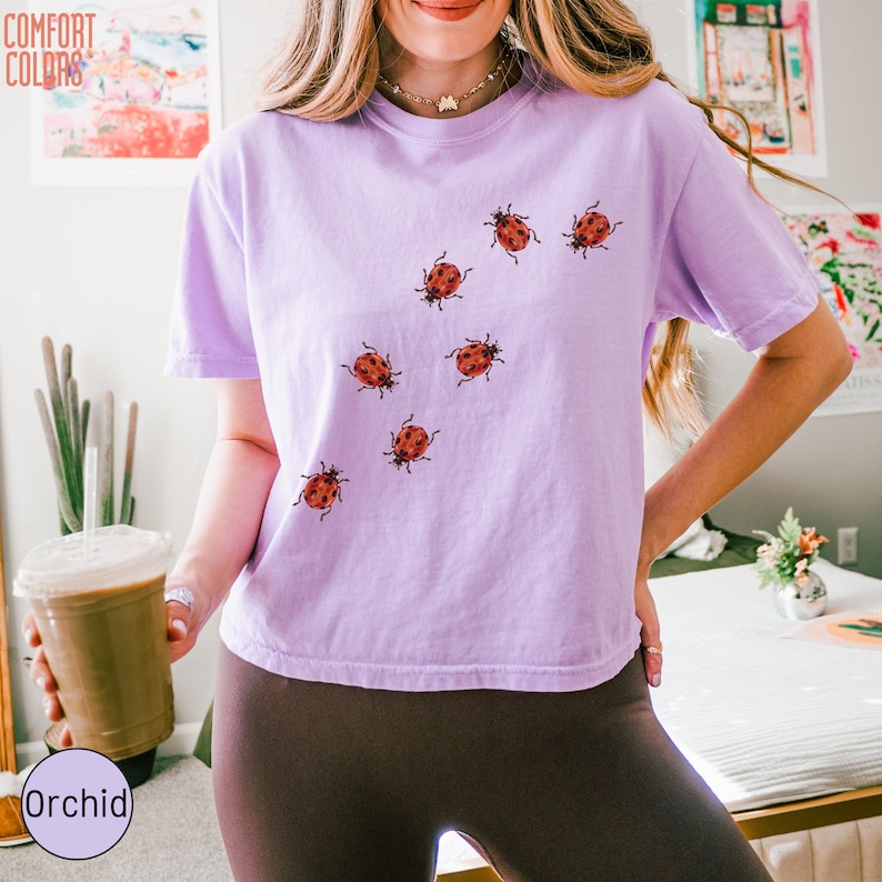 May include: A light purple short-sleeve t-shirt featuring a scattered pattern of small red ladybugs across the front. The shirt has a crew neckline and a relaxed fit. The text COMFORT COLORS is visible in the upper left corner of the image.