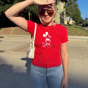 May include: A red t-shirt featuring a white graphic of Mickey Mouse. The shirt is short-sleeved and worn with light blue jeans and a white purse. The person is wearing sunglasses and is outside on a sunny day.