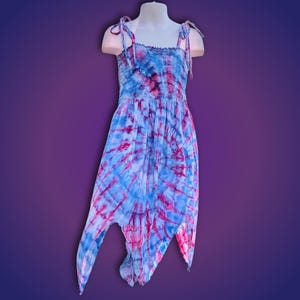 Puede incluir: Vestido tie-dye azul y rosa sobre maniquí. El vestido tiene un corpiño fruncido y tirantes anudados. La falda tiene un dobladillo asimétrico, creando una silueta fluida. La tela tiene un patrón arremolinado. Un vestido de verano.