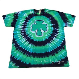 May include: A green, blue, and white tie-dye t-shirt with a green shamrock design in the center. The shirt has the text "Vivid Art by Regina" at the bottom.