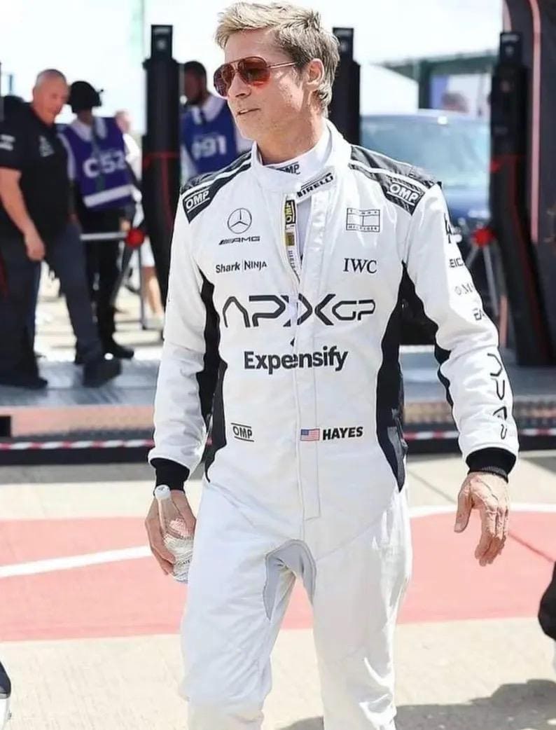 Puede incluir: Un mono de carreras blanco con detalles en negro, con logotipos de patrocinadores como "APXGP" y "Expensify". El traje incluye la bandera estadounidense y el nombre "HAYES". La persona lleva gafas de sol y sostiene una botella de agua.