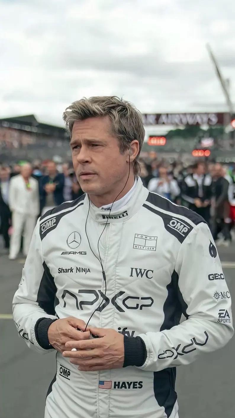 Puede incluir: Una chaqueta de carreras blanca con detalles en negro, con logotipos de patrocinadores como "OMP," "AMG" e "IWC." La chaqueta presenta "Shark NINJA" y "HAYES" con una bandera estadounidense. La persona lleva auriculares.