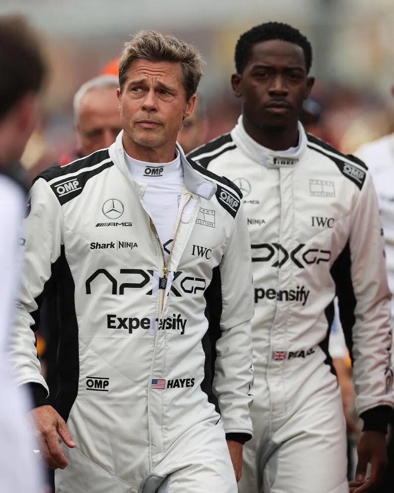 Puede incluir: Dos personas con trajes de carreras blancos con detalles negros y logotipos de patrocinadores. Los trajes tienen los logotipos de AMG, Shark Ninja, IWC y APXGP. Un traje tiene un peque&ntilde;o parche con la bandera estadounidense. Los trajes est&aacute;n cerrados con cremallera y tienen logotipos de OMP.