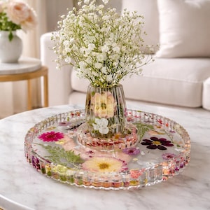 May include: A clear glass vase with a bouquet of white flowers sits on a round tray. The tray is filled with colorful dried flowers and greenery, set on a marble-topped coffee table with a gold base.