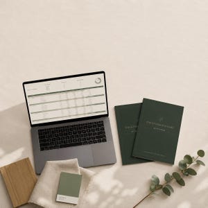 May include: A silver laptop displaying a spreadsheet, two dark green planners titled The Considered Home, a small sage green paint swatch, a folded beige fabric, and a wooden board next to a green eucalyptus branch with small round leaves.