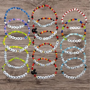 Könnte beinhalten: Eine Sammlung von bunten Armbändern mit Perlen, die Wörter wie "Run Disney" und "Magical" tragen. Einige Armbänder haben Mickey Mouse-Charms. Die Armbänder liegen auf einer Holzoberfläche.
