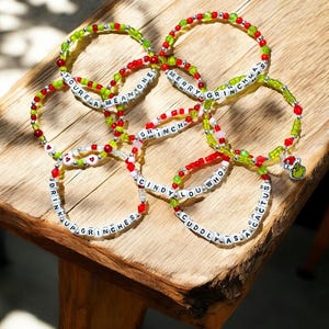 May include: Several beaded bracelets with festive Christmas messages. The bracelets are made with red, green, and white beads, with white letter beads forming phrases such as "MERRY GRINCHMAS" and "CINDY LOU WHO". Some bracelets have Grinch charms.