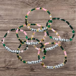 Puede incluir: Una colección de pulseras de cuentas en rosa, verde, dorado y blanco. Algunas pulseras deletrean "FOR GOOD", "WICKED", "ELPHABA" y "GLINDA". Cada pulsera presenta dijes en forma de estrella.