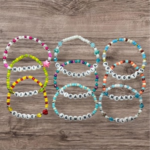 Può includere: Una collezione di braccialetti di perline con perline di lettere bianche che compongono nomi come "Minnie", "Cinderella" e "Pocahontas". I braccialetti presentano vari colori, tra cui rosa, verde, blu e arancione, con perline decorative.