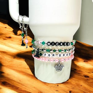 May include: A white tumbler with multiple beaded bracelets. The bracelets feature the words "WICKED" and "FOR GOOD" spelled out with letter beads. The bracelets are accented with green and pink beads, and charms including a witch hat and a crown.