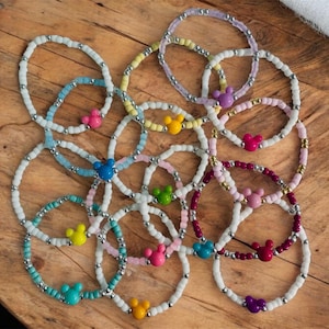 Puede incluir: Una colección de pulseras de cuentas en varios colores, incluyendo azul, rosa, amarillo y morado. Cada pulsera presenta un dije en forma de Mickey Mouse. Las pulseras se exhiben sobre una superficie de madera.