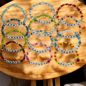 Könnte beinhalten: Eine Sammlung von Armbändern mit bunten Perlen und Buchstabenperlen, die Namen wie Jasmine, Elsa und Schneewittchen buchstabieren. Die Armbänder liegen auf einer Holzoberfläche.
