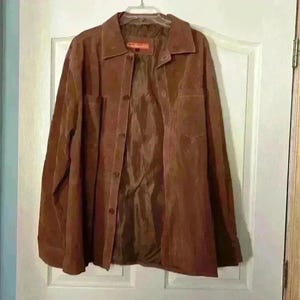 May include: A brown suede jacket with a button-up front and a collared neckline. The jacket has two front pockets and is hanging on a white door. The jacket is a medium brown color.