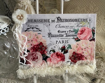 May include: A white canvas sign with a floral design and the text "Chemiserie du Patronometre" and "Chemises sur Mesure". The sign is decorated with a white lace trim and a white flower with pearl accents.