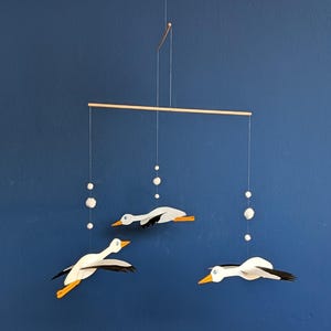 Op de afbeelding: Een houten mobiel met drie witte en zwarte vogels met oranje snavels en poten, hangend aan een houten balk. De mobiel bevat witte pompons en staat tegen een diepblauwe achtergrond.