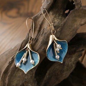 May include: A pair of gold-tone earrings featuring a blue enamel lily pad design with gold accents and faux pearls. The earrings have a long hook style for pierced ears. The earrings are displayed on a piece of driftwood.