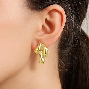 Può includere: Un paio di orecchini a lobo dorati dal design astratto. Gli orecchini presentano un design fluido e organico con tre elementi collegati a forma di goccia. Gli orecchini sono mostrati su un orecchio.