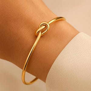 Puede incluir: Una pulsera dorada con un diseño de nudo. La pulsera está hecha de un metal brillante y liso y se lleva en la muñeca. El nudo es el punto focal, agregando un toque único al diseño simple. Una pieza de joyería clásica.