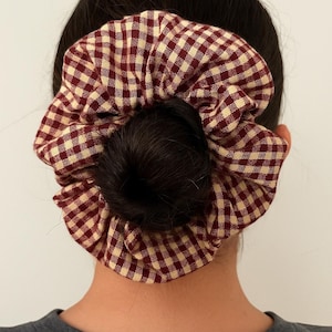 May include: A burgundy and cream gingham scrunchie encircles a dark bun. The hair accessory is made of a soft, textured fabric. The scrunchie is a hair accessory, perfect for adding a touch of style to any hairstyle.