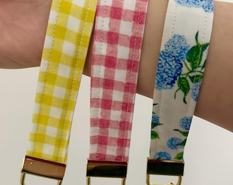 May include: Three fabric keychains in yellow gingham, pink gingham, and white with blue floral patterns. Each keychain has a gold-colored metal clasp and key ring. The keychains are displayed on a wrist.