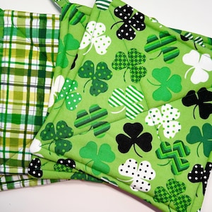 May include: A stack of three square pot holders. The pot holders feature green and white plaid and shamrock patterns. The shamrock patterns include white polka dots, black polka dots, and green and white stripes. Each pot holder has a fabric loop.