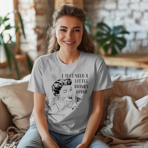May include: A light gray t-shirt featuring the text "I JUST NEED A LITTLE DRINKY DRINK" with a black and white illustration of a woman holding a drink. The shirt is a crew neck style.