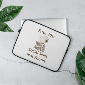 May include: White laptop sleeve with a black zipper and trim. The sleeve features a cartoon robot reading a book, with the text "Error 404: Social Skills Not Found."