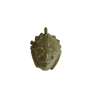 May include: A small, green bronze sculpture of a face with a detailed design. The sculpture is a pendant and has a loop at the top for hanging.