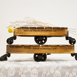 May include: Two wooden crates with wheels, stacked on top of each other. The top crate has dried flowers and a yellow pom-pom. Both crates have the text "OLD NEW HOUSE, SCARSDALE, N.Y."