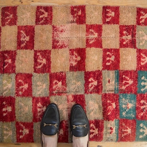 May include: A red and beige checkered rug with a pattern of stylized figures. Two black leather loafers with gold buckles are standing on the rug.