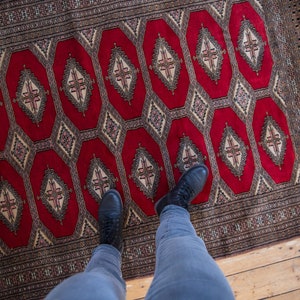 May include: A person wearing blue jeans and black shoes stands on a red and brown patterned rug. The rug has a geometric design with repeating diamond shapes.
