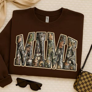 May include: A brown sweatshirt with the word "MAMA" in large, block letters across the front. The letters are filled with a camouflage pattern and outlined in a cream color. The sweatshirt has a crew neck and long sleeves.