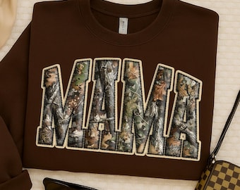 May include: A brown sweatshirt with the word "MAMA" in large, block letters across the front. The letters are filled with a camouflage pattern and outlined in a cream color. The sweatshirt has a crew neck and long sleeves.