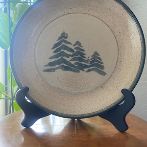 May include: A ceramic plate with a beige center and a dark green rim. The plate features a blue-green illustration of three stylized pine trees. The plate is displayed on a black stand.