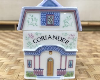 May include: A white ceramic spice jar shaped like a house, labeled "Coriander" in script. The house has a blue roof, pink accents, and a small porch. Floral and leaf designs decorate the exterior.