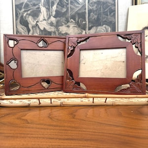 May include: Two wooden picture frames with a reddish-brown finish. One frame features heart-shaped cutouts, while the other has leaf-shaped carvings. Both frames have a rectangular opening for photos.