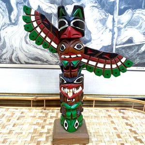 May include: A hand-carved wooden totem pole with multiple figures, featuring a bird with outstretched wings. The totem pole is painted in vibrant colors, including red, green, black, and brown. It stands on a small, rectangular wooden base.