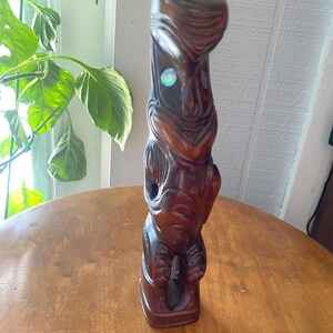 May include: A dark brown, carved wooden tiki statue with a rectangular base. The statue features a detailed face with a small, iridescent inlay. The tiki is standing on a round, wooden table.