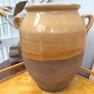 May include: A large, two-handled ceramic jar with a wide mouth. The upper portion is a light tan color, transitioning to a darker brown at the base. The jar sits on a woven tray.