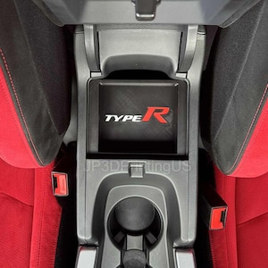 May include: Interior car detail featuring a center console with a black storage compartment. The compartment has the text "TYPE R" in white and red. Red and black seats surround the console, with red seatbelts visible.